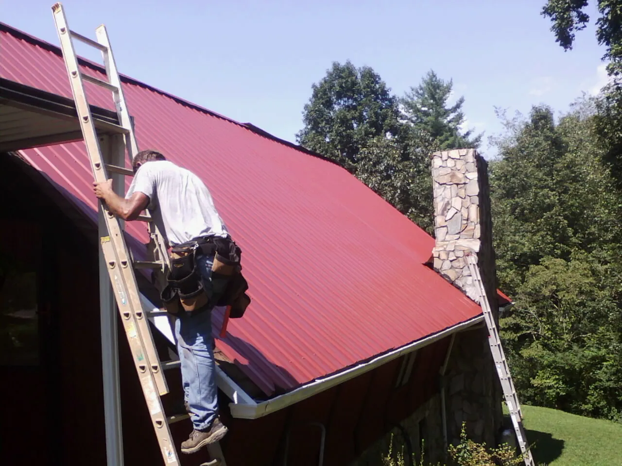 Roofer on a ladder beside a red metal roof and stone chimney — Soffit & Fascia Repair by Roofing Brothers in Flemingsburg